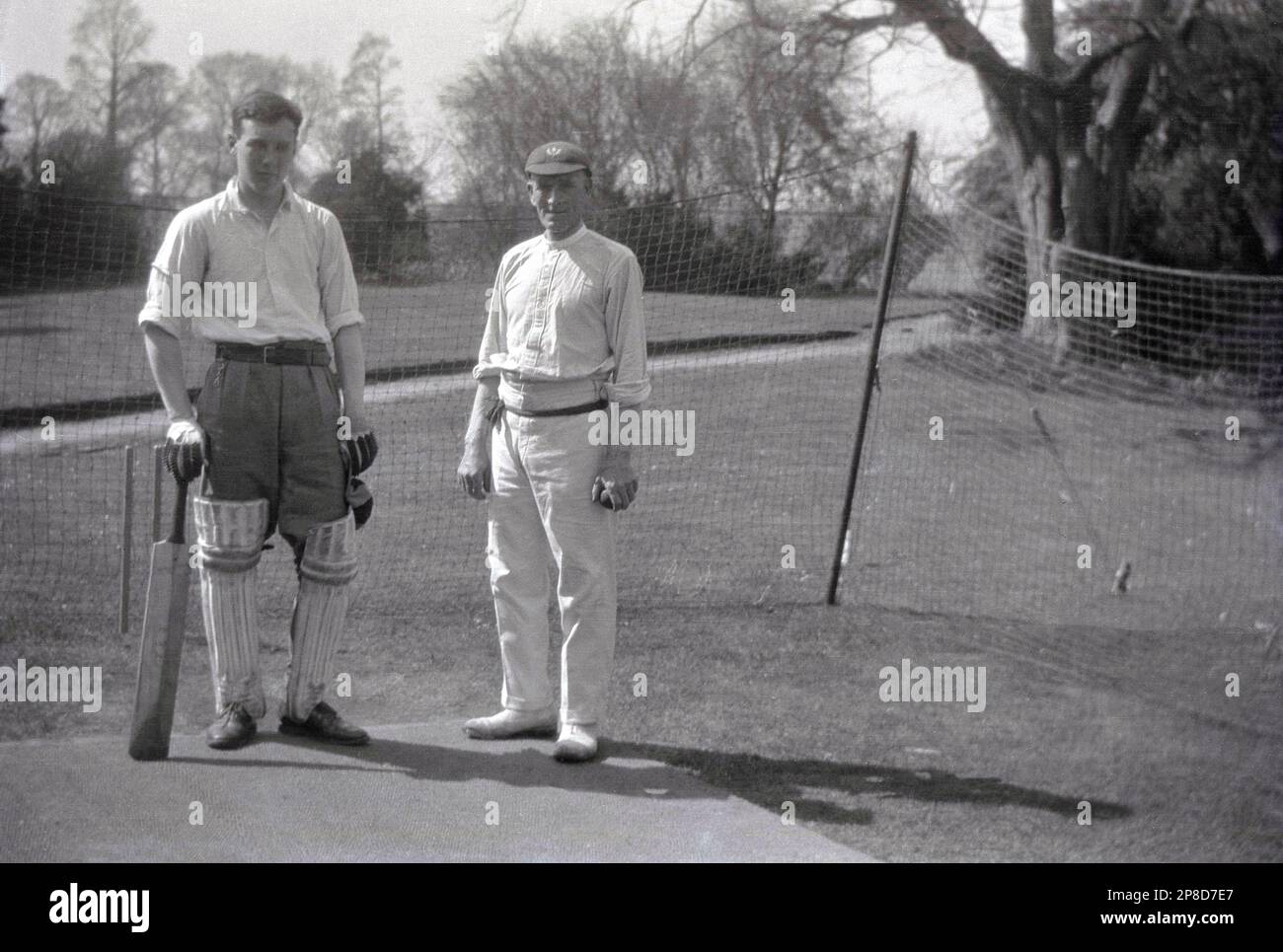 Circa 1930s, storico, un giovane uomo che indossa i pattini, i guanti e che tiene un pipistrello che ha una rete di cricket su un prato nel terreno di una casa di campagna, Inghilterra, Regno Unito. Sta in piedi per la sua foto accanto a un più grande grillo maschio adulto, il bowler, che sta portando i bianchi del cricket del giorno, pantaloni o flanelle sostenuti da cravatta della vita, camicia del collo rotondo e cappuccio. Foto Stock