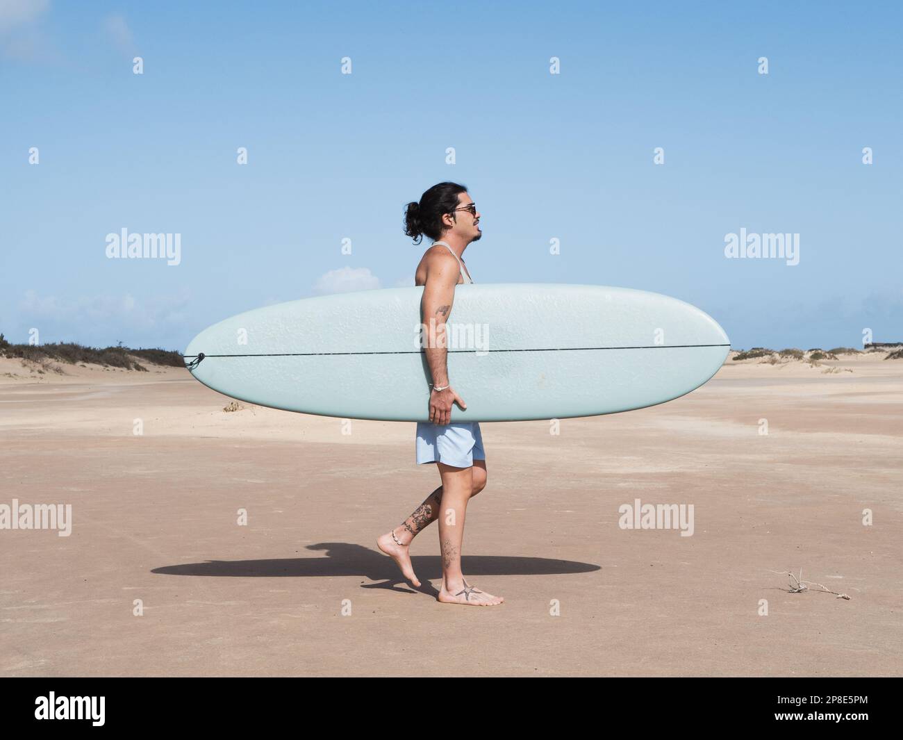 Surfista ispanico che cammina sulla sabbia tenendo la tavola da surf Foto Stock
