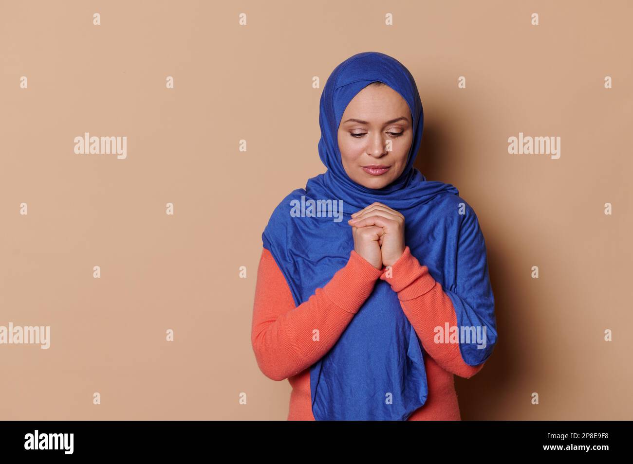 Donna musulmana in hijab blu, tenendo le mani piegate al livello del petto, pregando per la pace, isolata su sfondo beige Foto Stock