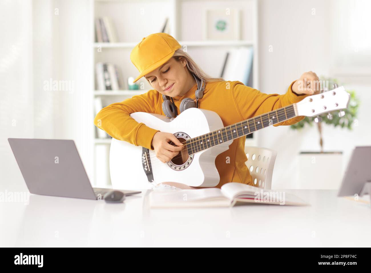 Giovane donna che sintonizza una chitarra acustica di fronte a un computer portatile a casa Foto Stock