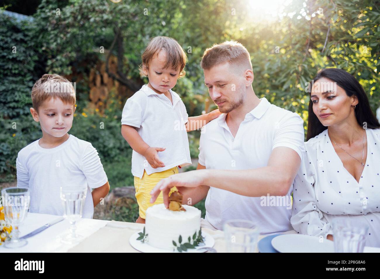 Felice famiglia bianca celebrare il compleanno. Mamma, papà e due piccoli figli in abiti bianchi mangiano una torta in un caffè all'aperto. Alberi verdi sullo sfondo. Copia Foto Stock