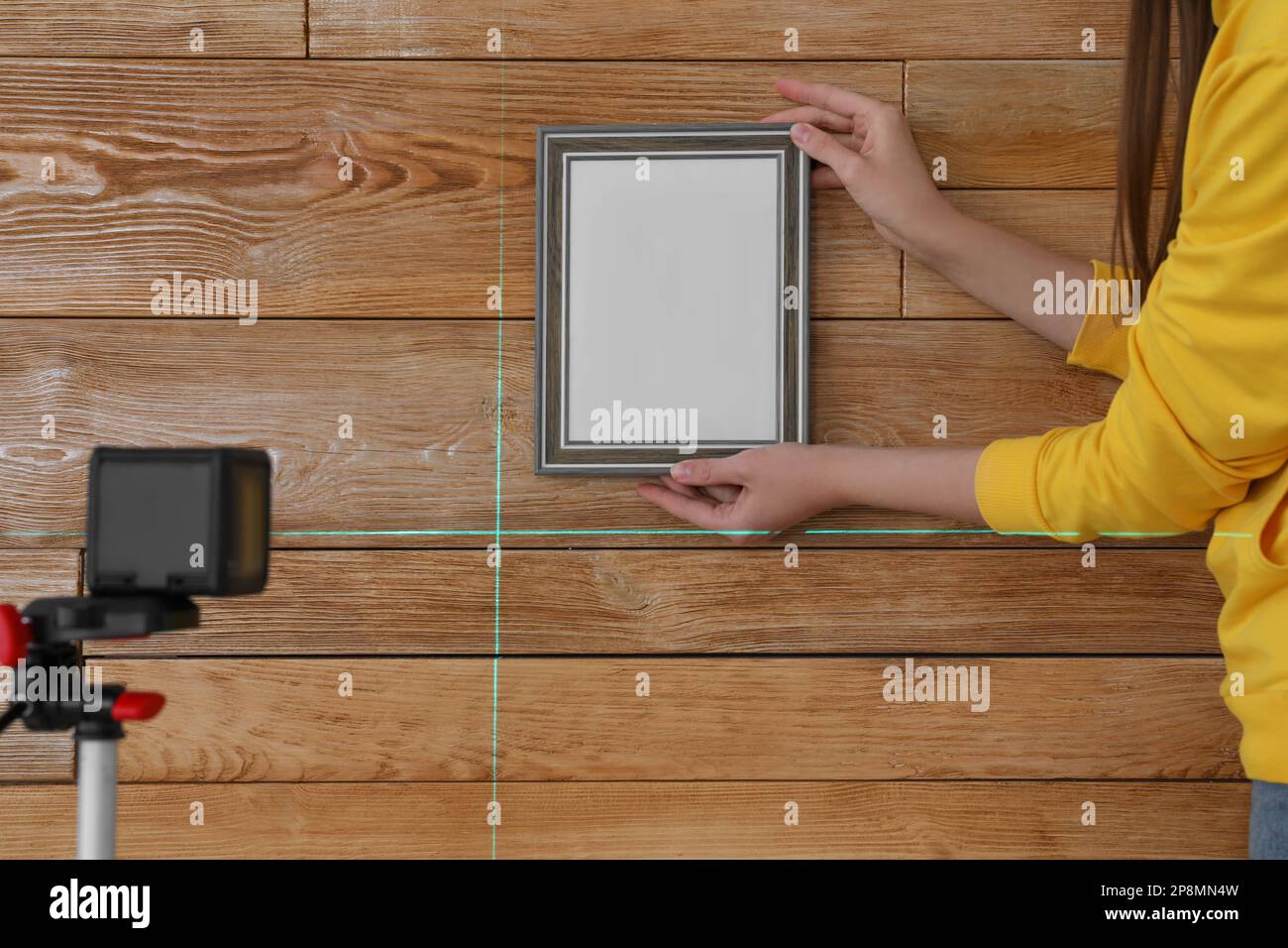 Donna che utilizza livella laser a linee incrociate per appendere una cornice fotografica vuota su una parete di legno, primo piano Foto Stock