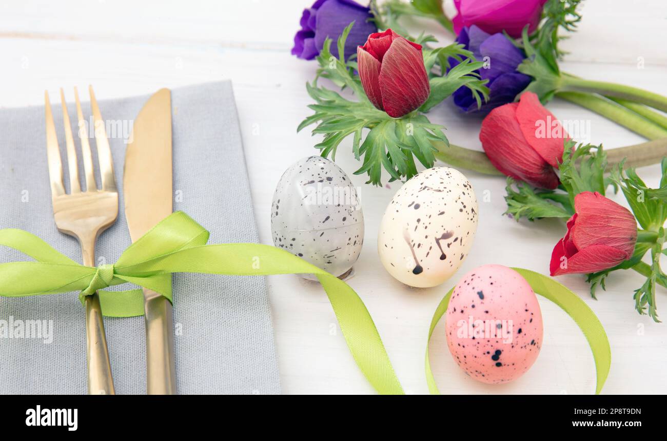 Tavola di Pasqua, fiori primaverili e decorazione uova, tovagliolo e posate dorate su sfondo di legno bianco Foto Stock