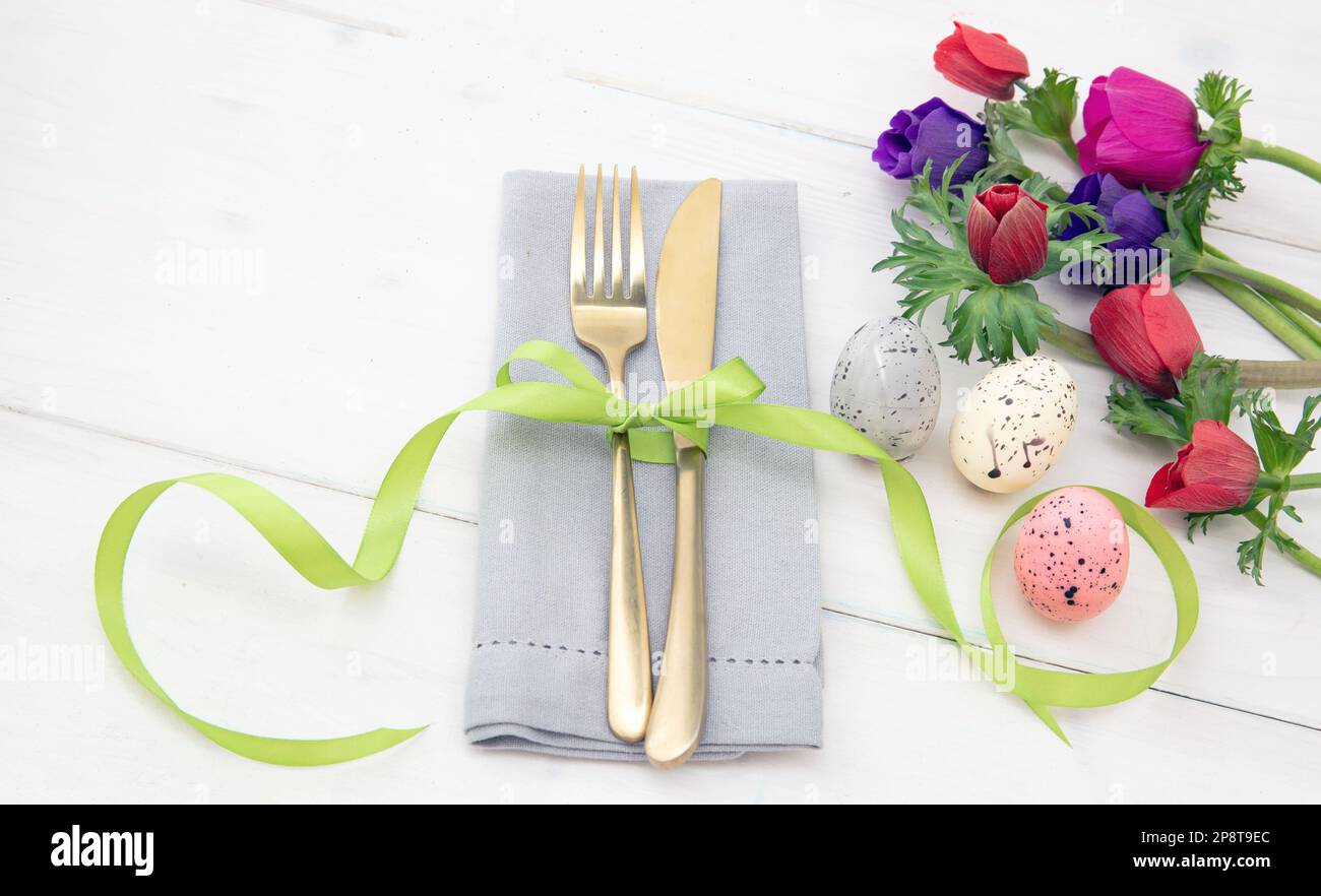 Tavola di Pasqua, fiori primaverili e decorazione uova, tovagliolo e posate dorate su sfondo di legno bianco Foto Stock