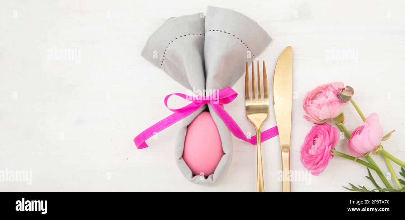 Tavolo di Pasqua, uovo di Pasqua rosa in un tovagliolo grigio, forma di orecchie di coniglio e posate dorate su sfondo di legno bianco, vista dall'alto Foto Stock