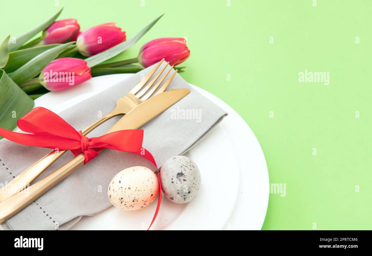 Tavola di Pasqua, fiori primaverili e uova decorazione, tovagliolo e posate dorate su piatti bianchi, sfondo verde pastello Foto Stock