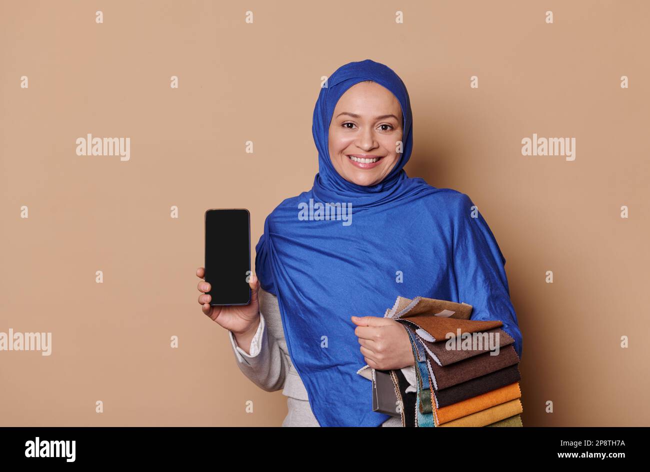 Bella donna musulmana in hijab blu, responsabile vendite in negozio di mobili, mostra uno smartphone con schermo vuoto per app mobile Foto Stock