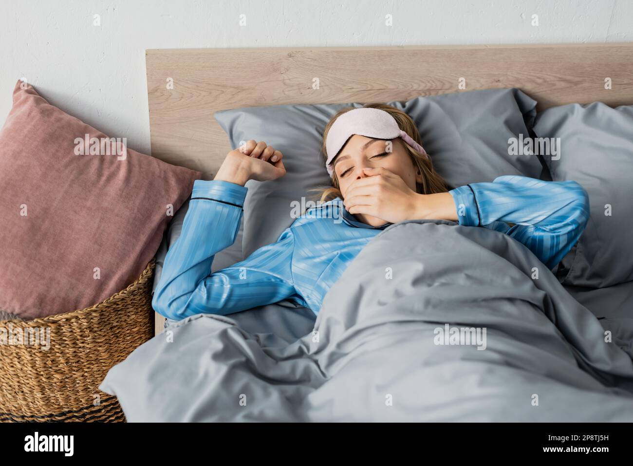 donna addormentata in pajama di seta e maschera notturna che sbadigna e copre la bocca mentre sdraiata a letto, immagine stock Foto Stock
