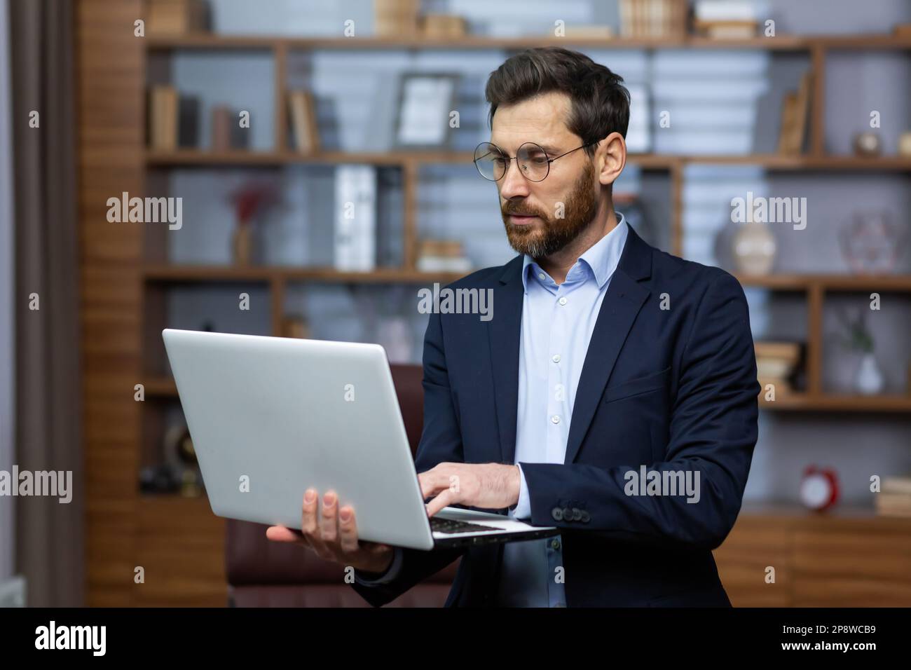 Serio giovane uomo d'affari avvocato in tuta e occhiali in piedi in ufficio e tenendo il computer portatile, al lavoro, digitando sulla tastiera. Foto Stock