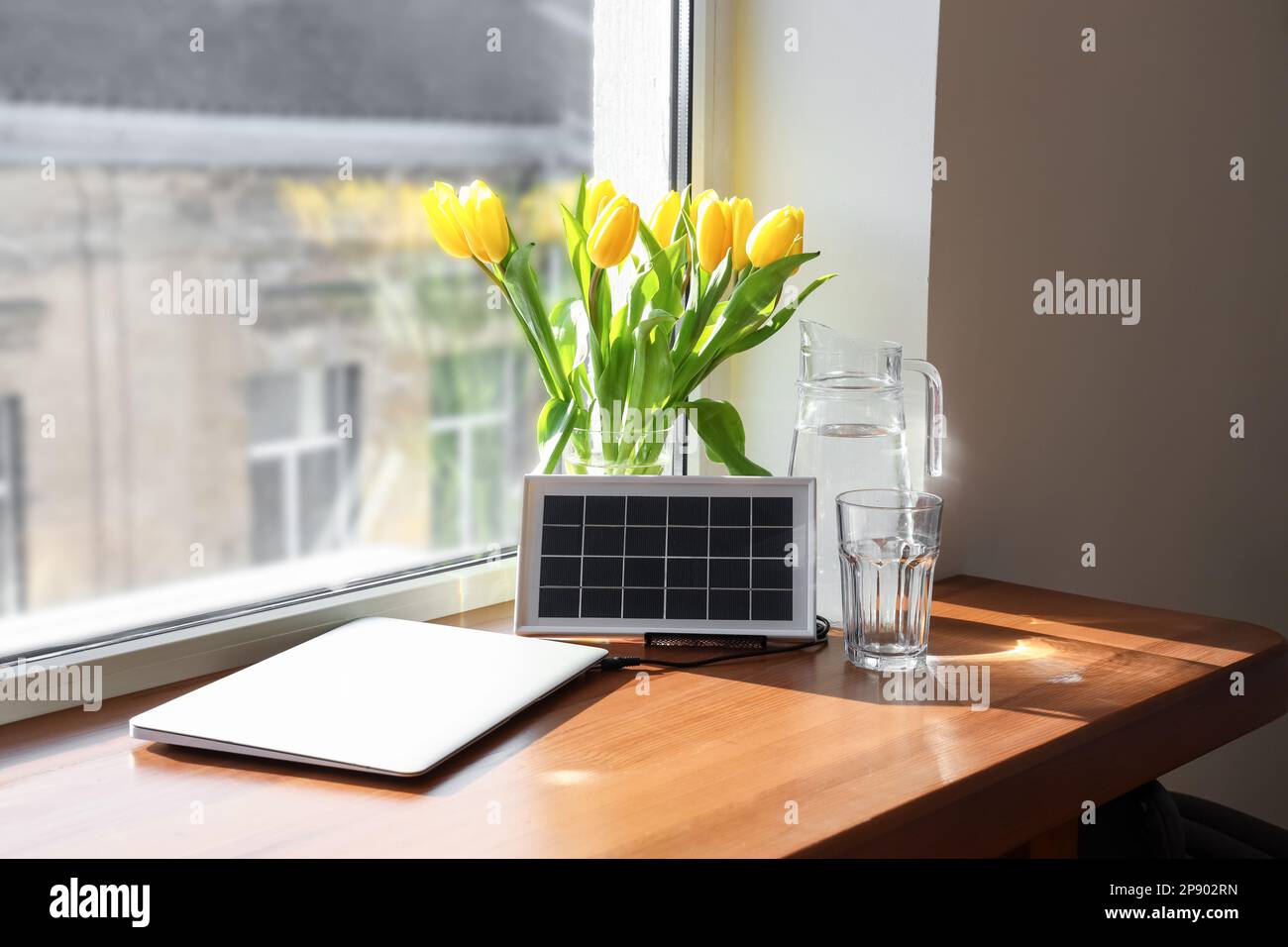 Portatile pannello solare di carica computer portatile, caraffa d'acqua e vaso con tulipani sul tavolo vicino alla finestra Foto Stock
