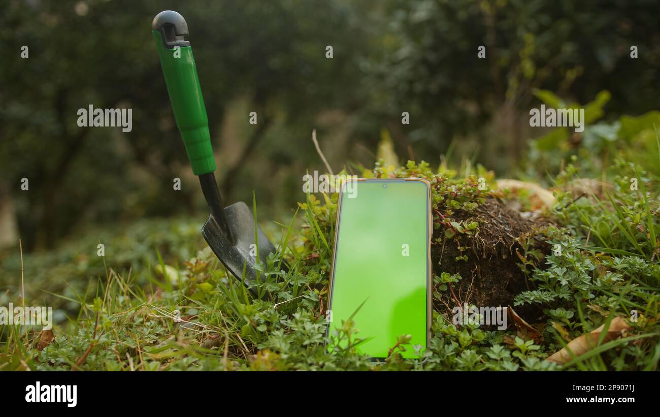Coltivazione digitale: Pala da giardiniere o piccola su erba con smartphone con schermo verde. Concetto di applicazione digitale e agricoltura futura Foto Stock