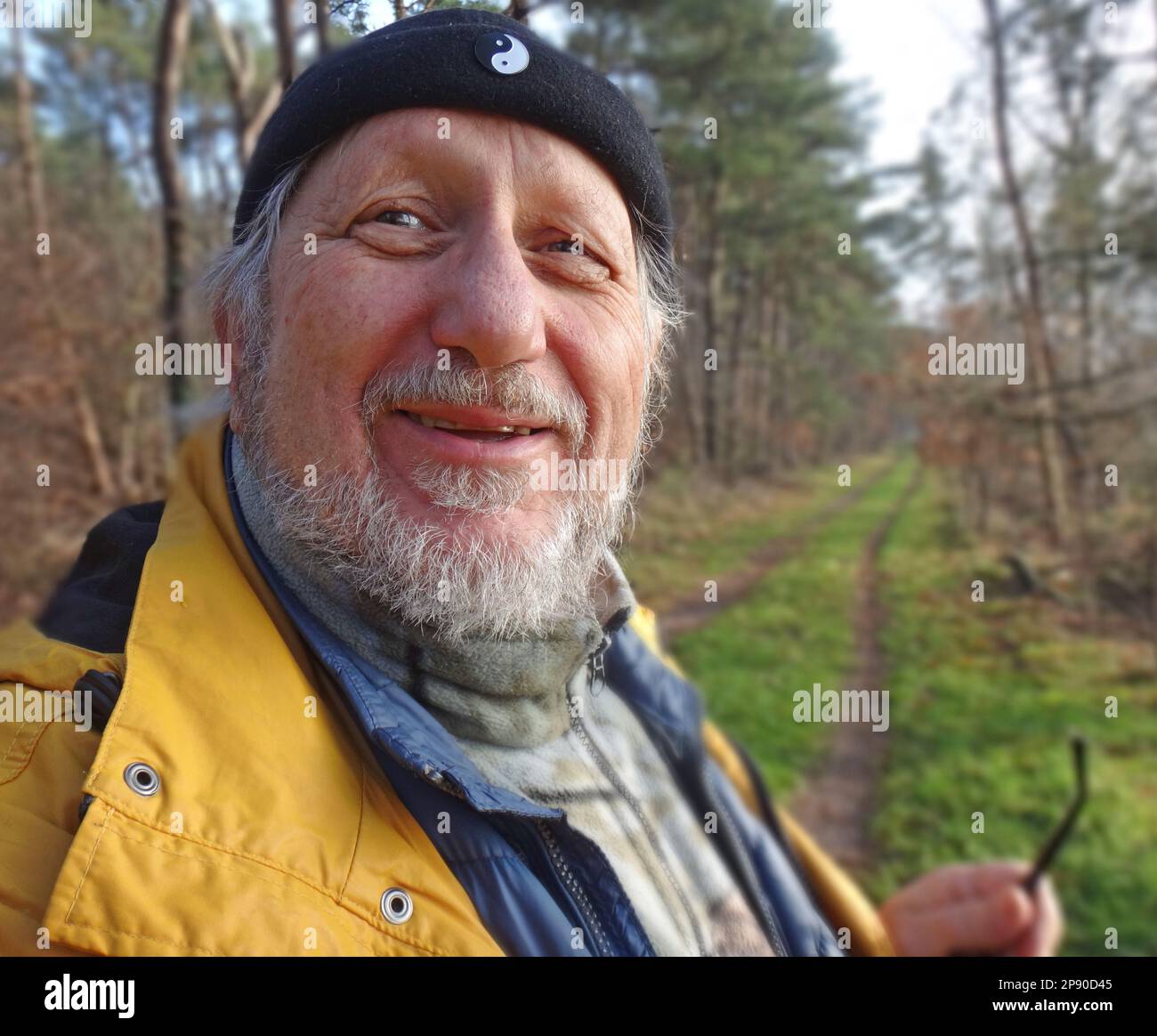 Amico uomo anziano con barba e berretto guardante dentro la macchina fotografica. Tolse gli occhiali. Si sta svegliando con un impermeabile giallo nel bosco. Foto Stock