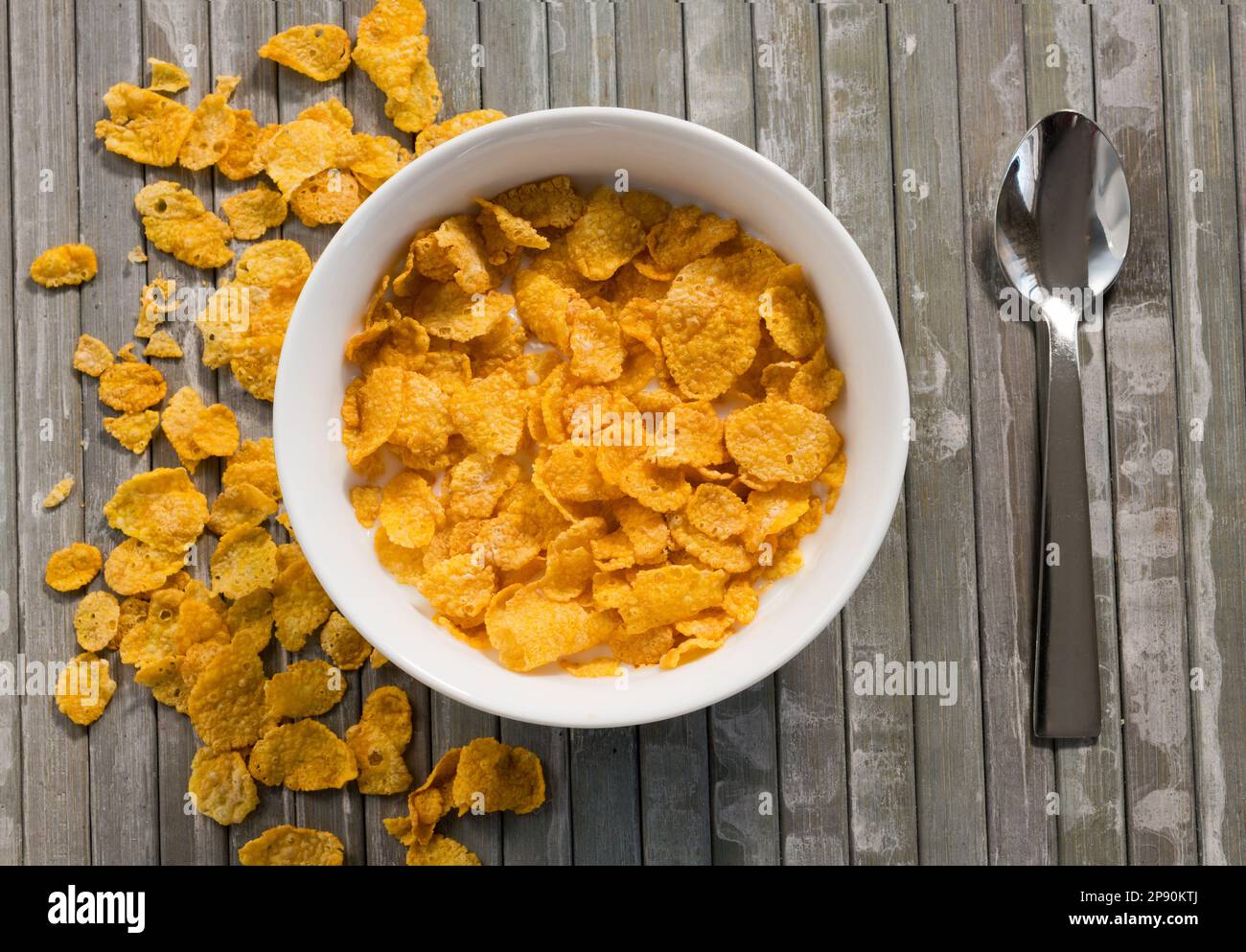 Vista dall'alto di appetitosi corn flakes in una ciotola bianca con latte posto su un tavolo di legno a strisce vicino al cucchiaio Foto Stock