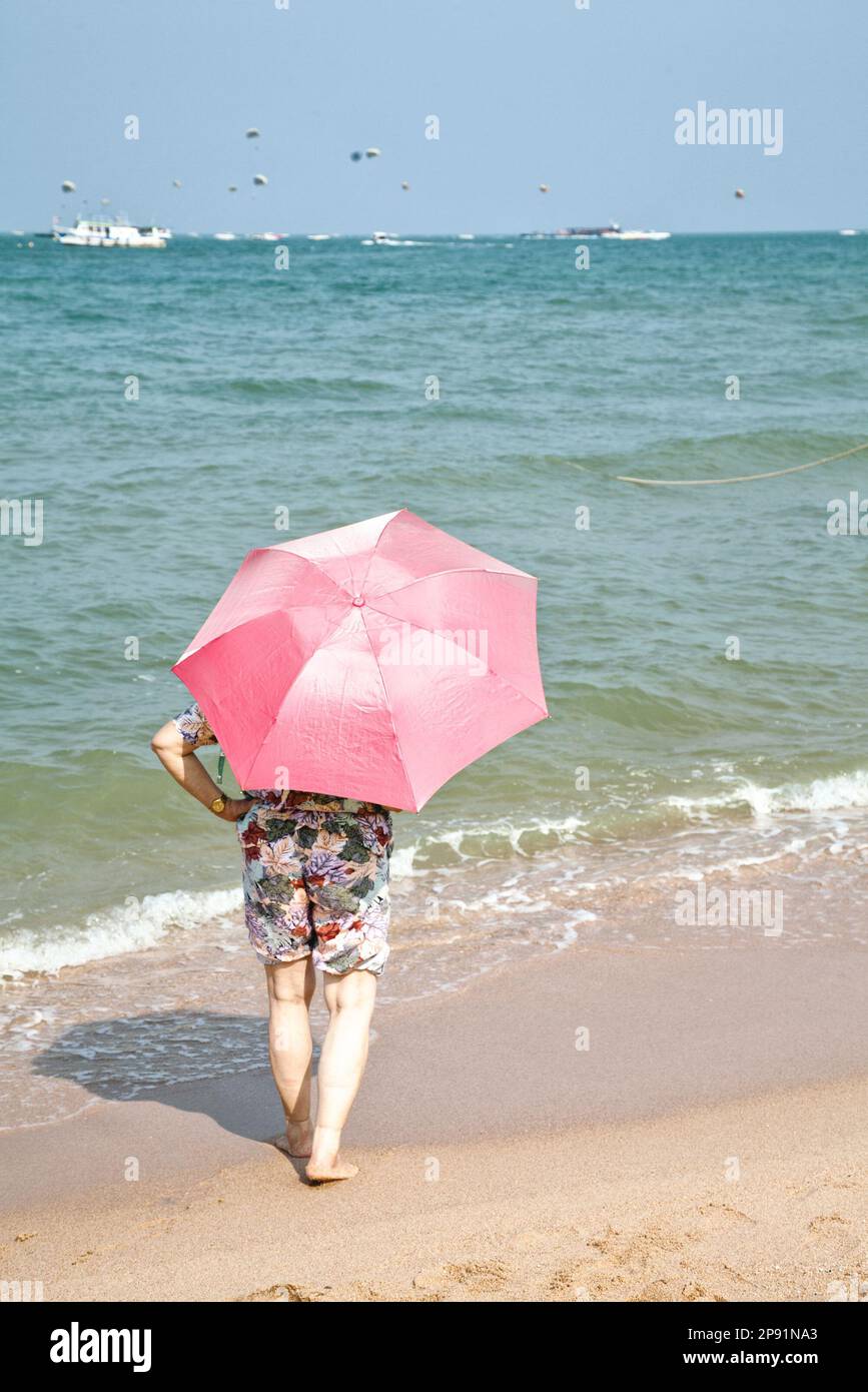 Donna anziana vestita di pantaloncini con un ombrello rosa in piedi su una spiaggia di sabbia, prendendo il sole, guardando all'orizzonte del mare Foto Stock