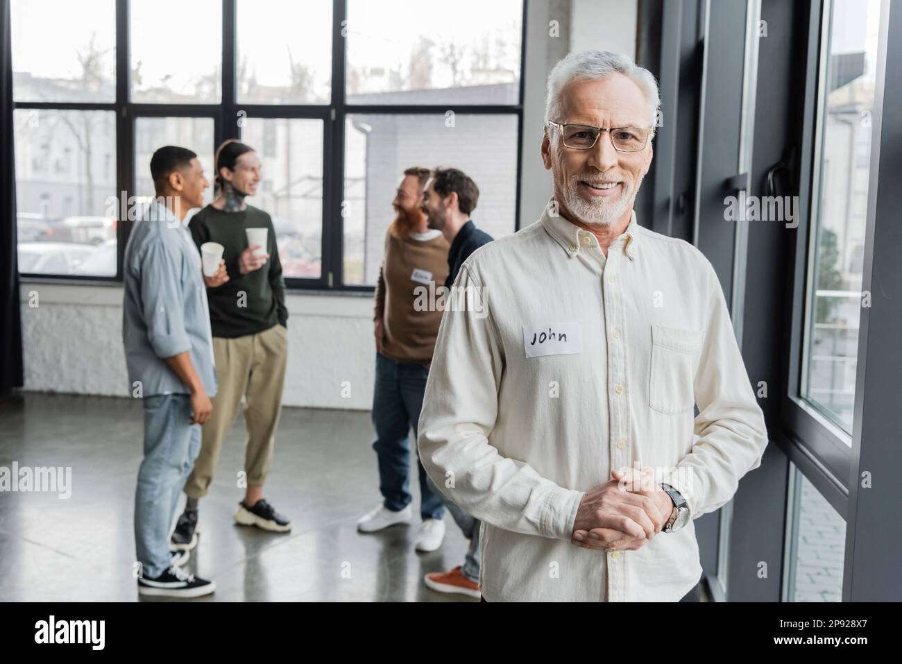Sorridente uomo di mezza età che guarda la macchina fotografica vicino a un gruppo sfocato durante la riunione degli alcolisti nel centro di riabilitazione, immagine di scorta Foto Stock