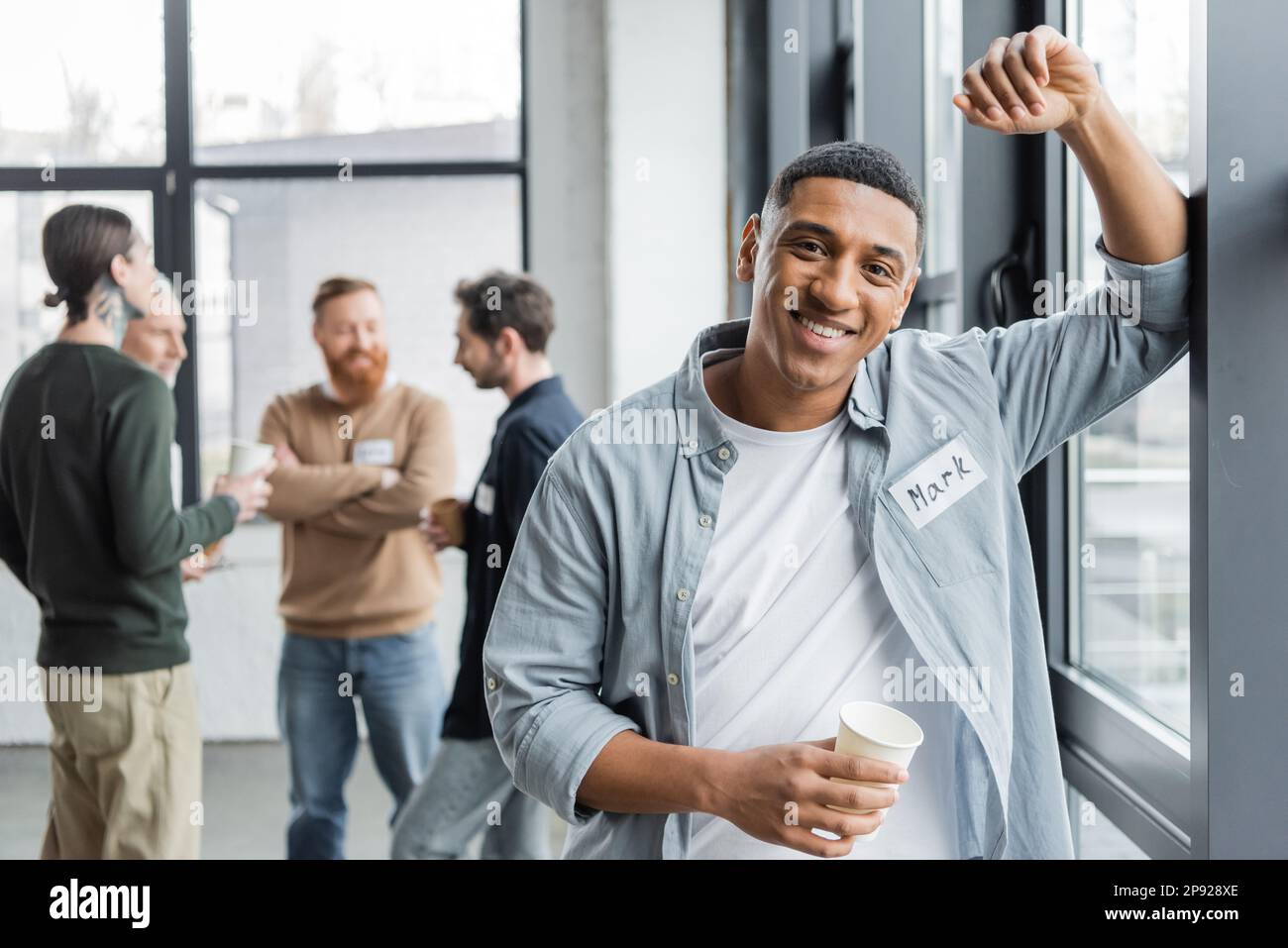 Uomo afroamericano sorridente che tiene la tazza di carta vicino al gruppo sfocato durante la riunione degli alcolisti nel centro di riabilitazione, immagine di scorta Foto Stock