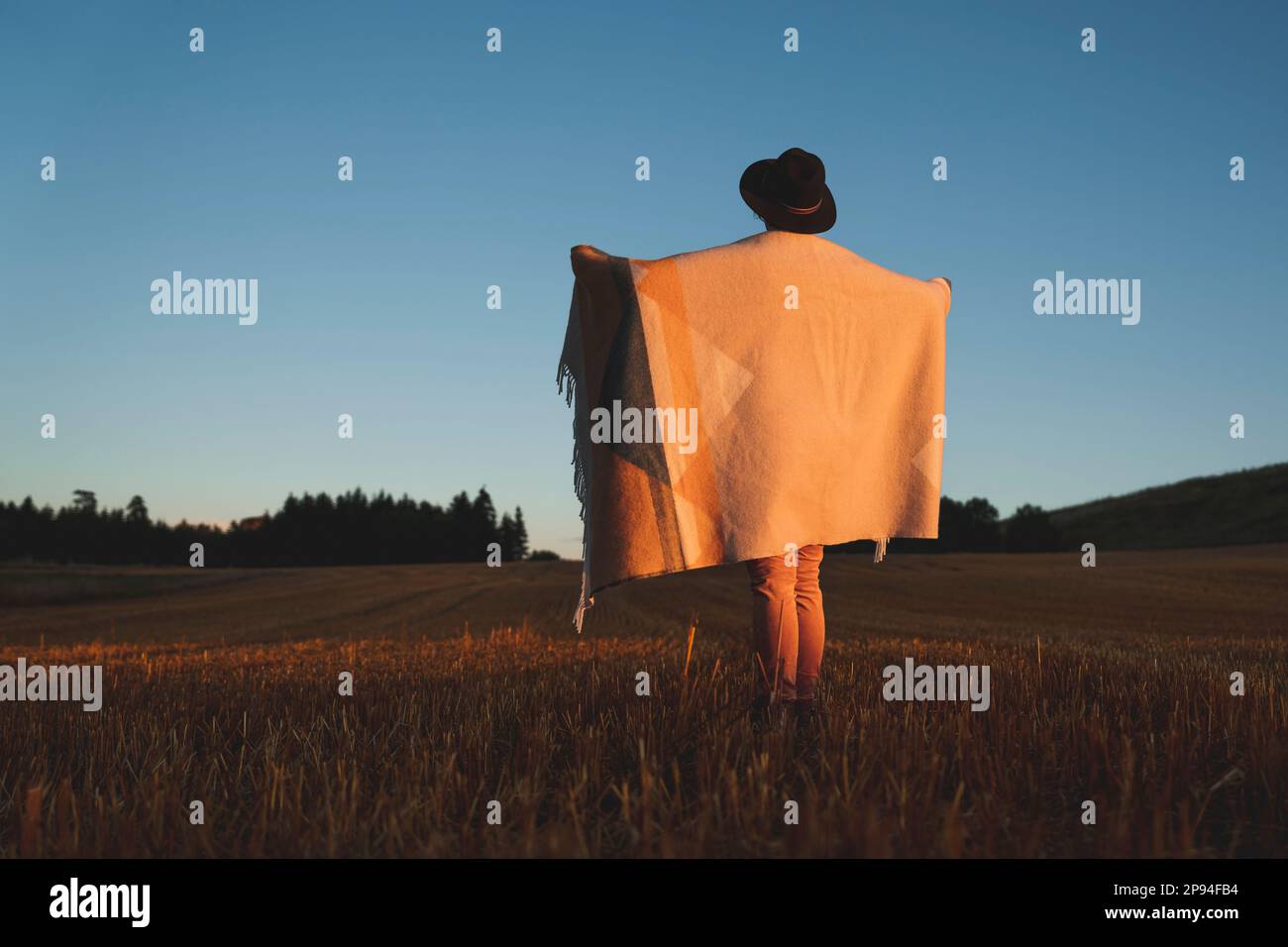 Donna con cappello e coperta di lana in campo stoppia, tramonto Foto Stock