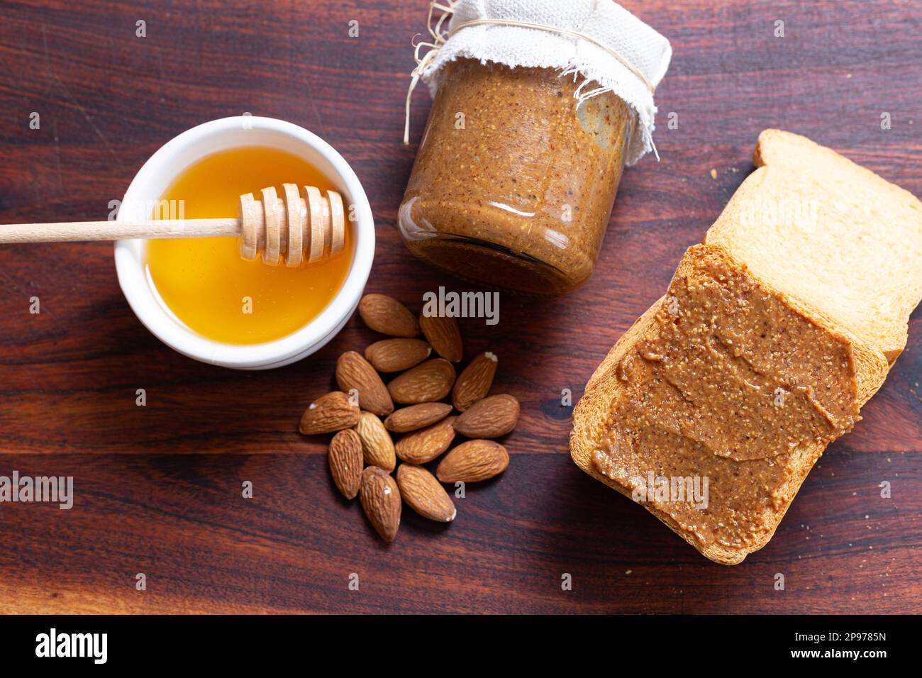 Sana colazione/spuntino: Miele, barattolo di burro di mandorle, mandorle, toast su legno rustico. Dolce e nutriente. Foto Stock