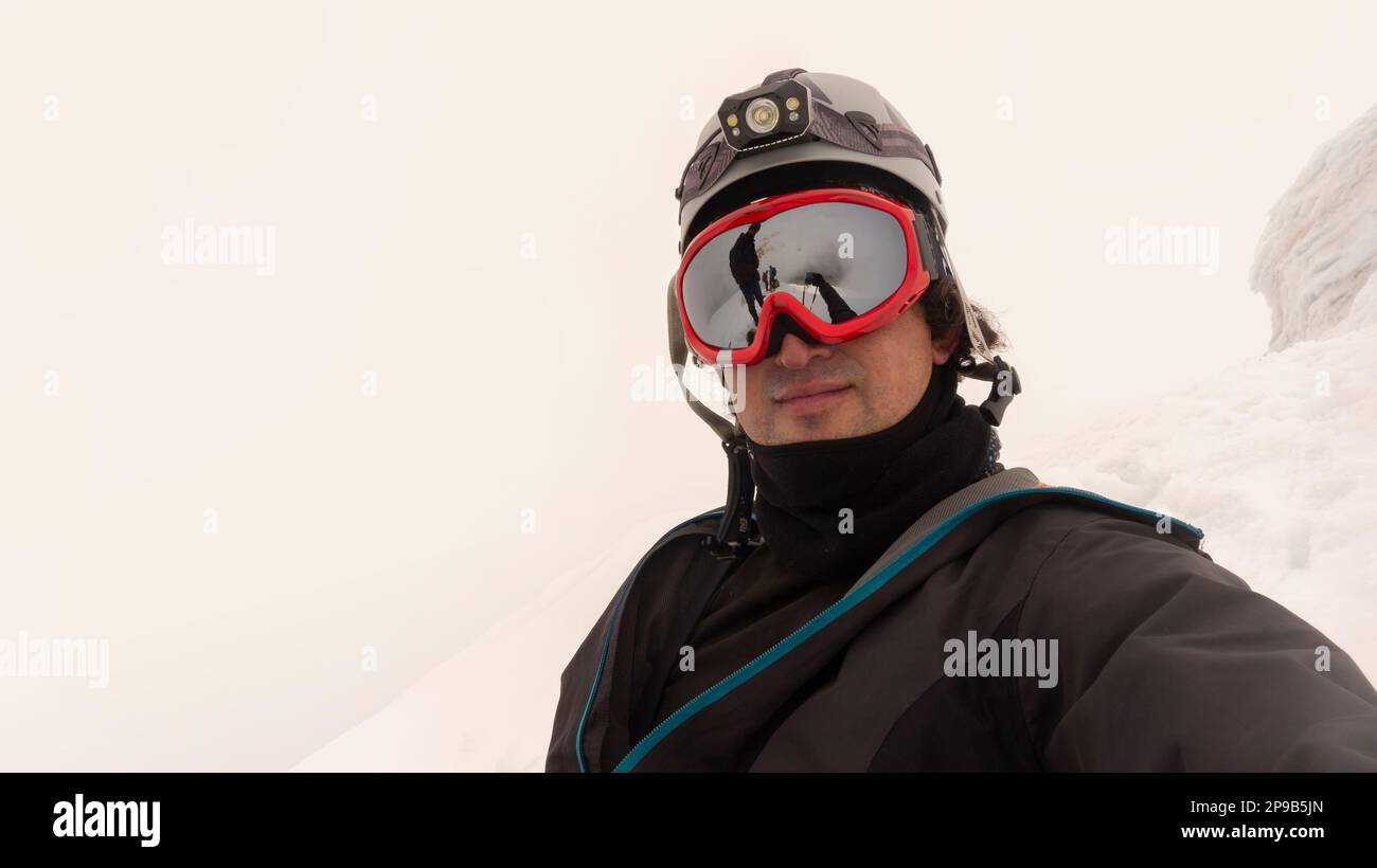 Arrampicatore con casco, faro e giacca nera che prende un selfie su un ghiacciaio in una giornata nuvolosa, il paesaggio si riflette in occhiali con cornice rossa Foto Stock