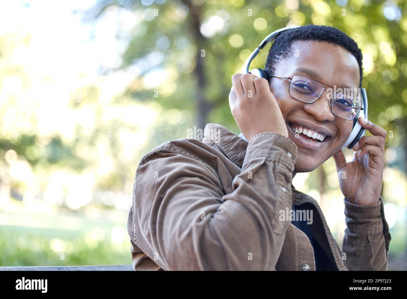 Cuffie, parco e ritratto dell'uomo africano che ascolta musica per esterni, salute mentale e rilassante pausa nella natura. Giovane, allievo felice o nero Foto Stock