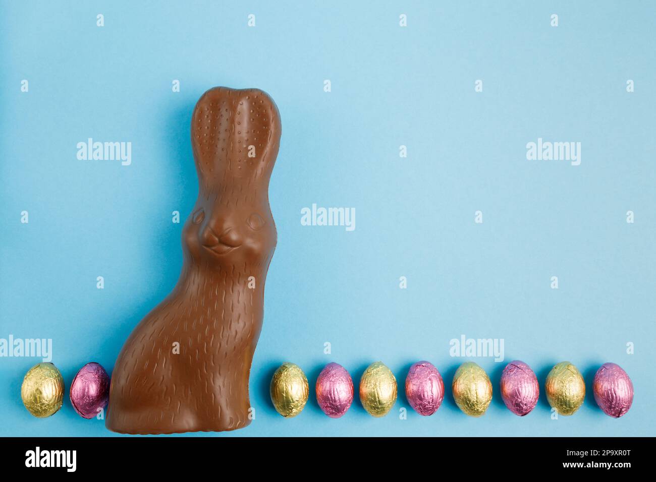 Coniglietto al cioccolato al latte e fila di uova di caramelle di Pasqua avvolte in foglio rosa e dorato su sfondo blu. Buon concetto di Pasqua. Preparazione per le vacanze. S Foto Stock