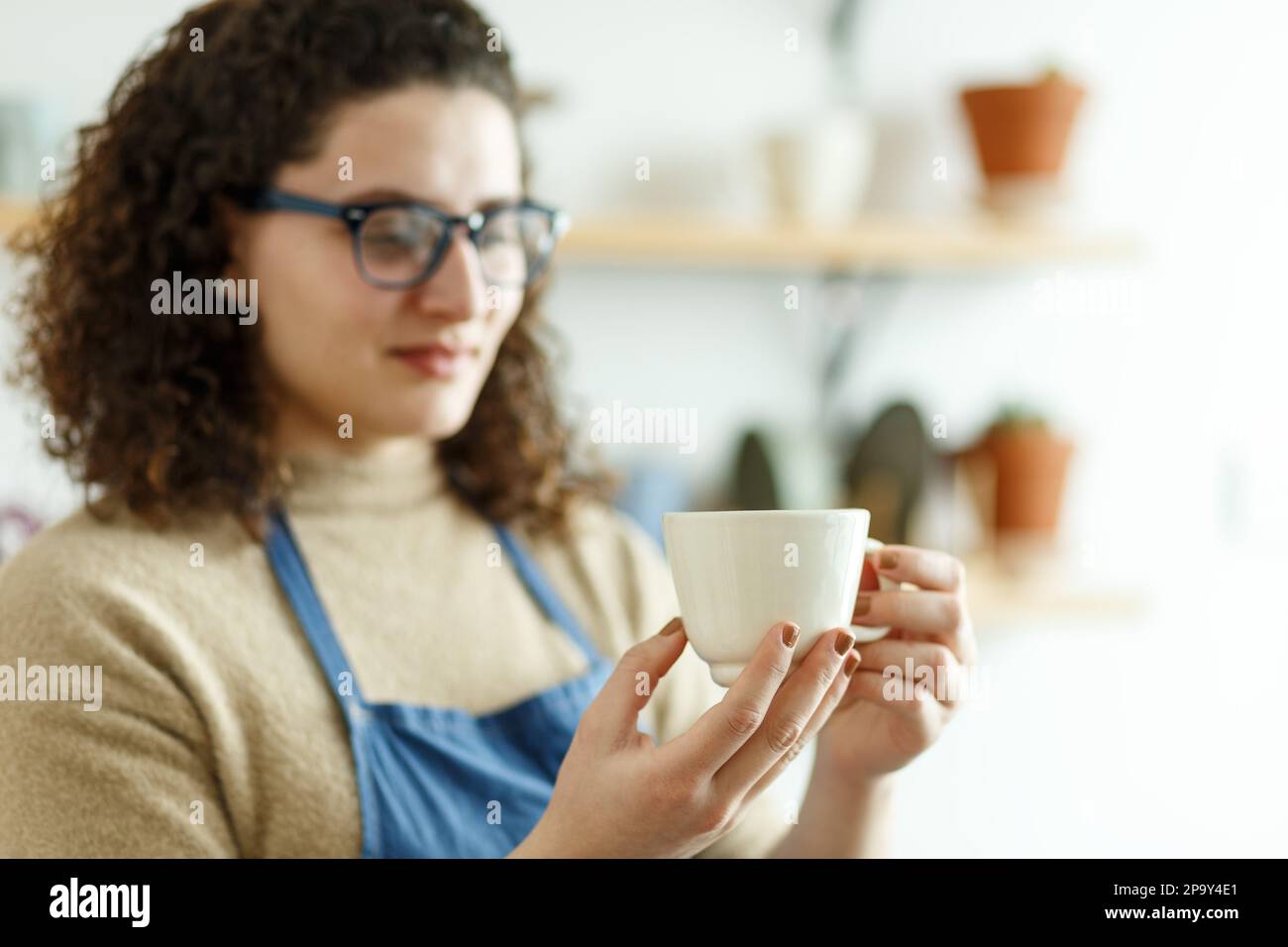 Felice giovane ceramista guardando via pensieroso mentre prende il calcio. Donna d'affari creativa che gestisce un negozio con prodotti in ceramica fatti a mano. Interno femminile Foto Stock