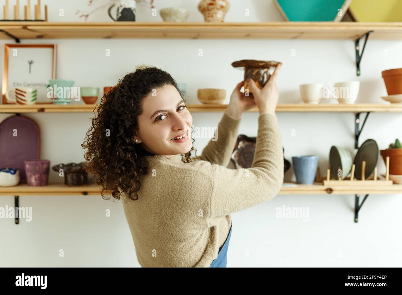 Felice giovane ceramista guardando via pensieroso mentre prende il calcio. Donna d'affari creativa che gestisce un negozio con prodotti in ceramica fatti a mano. Interno femminile Foto Stock