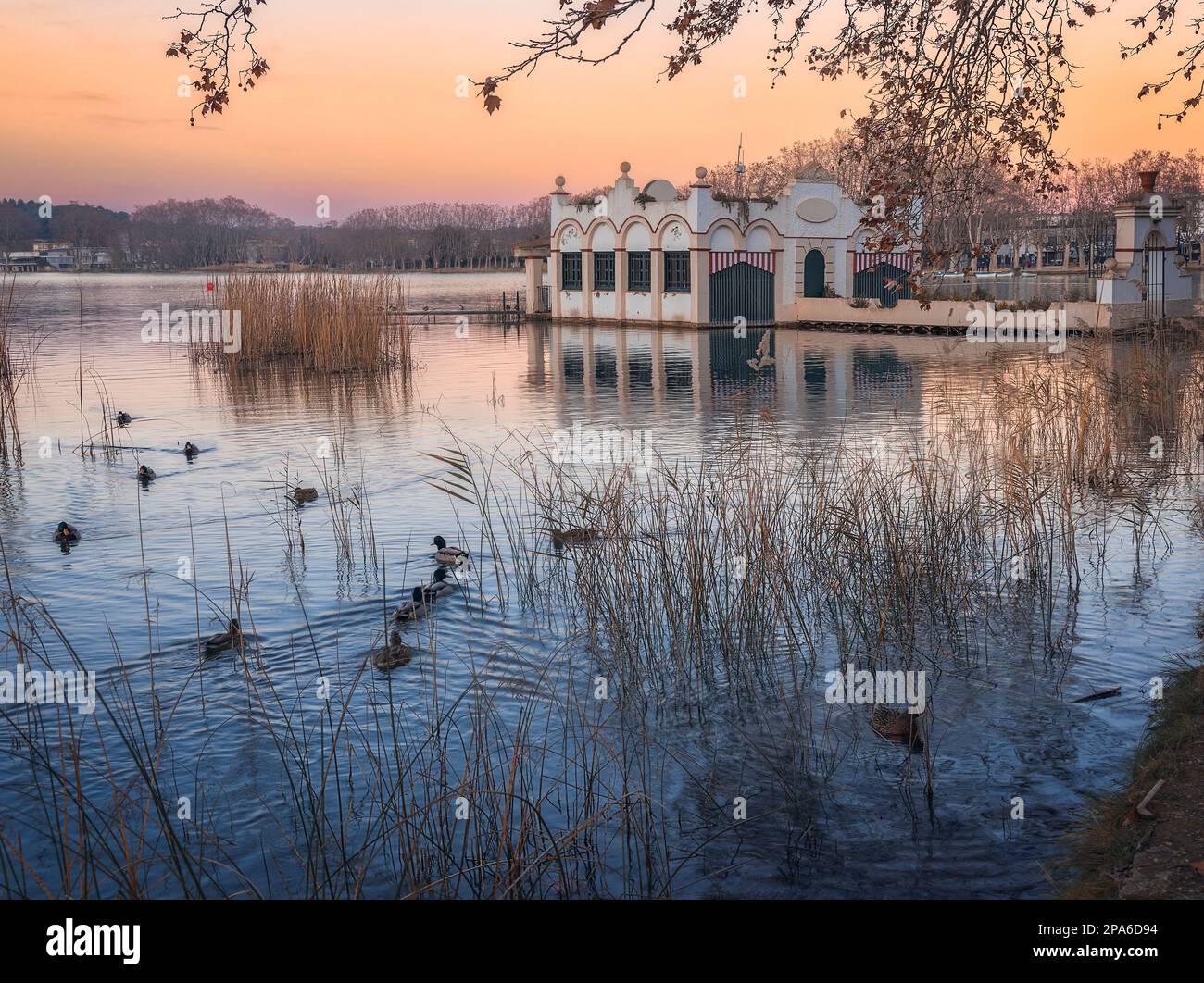 La tranquilla bellezza del lago Banyoles si riflette nel cielo al tramonto, incorniciato da una maestosa struttura costruita e circondato da alberi. Foto Stock