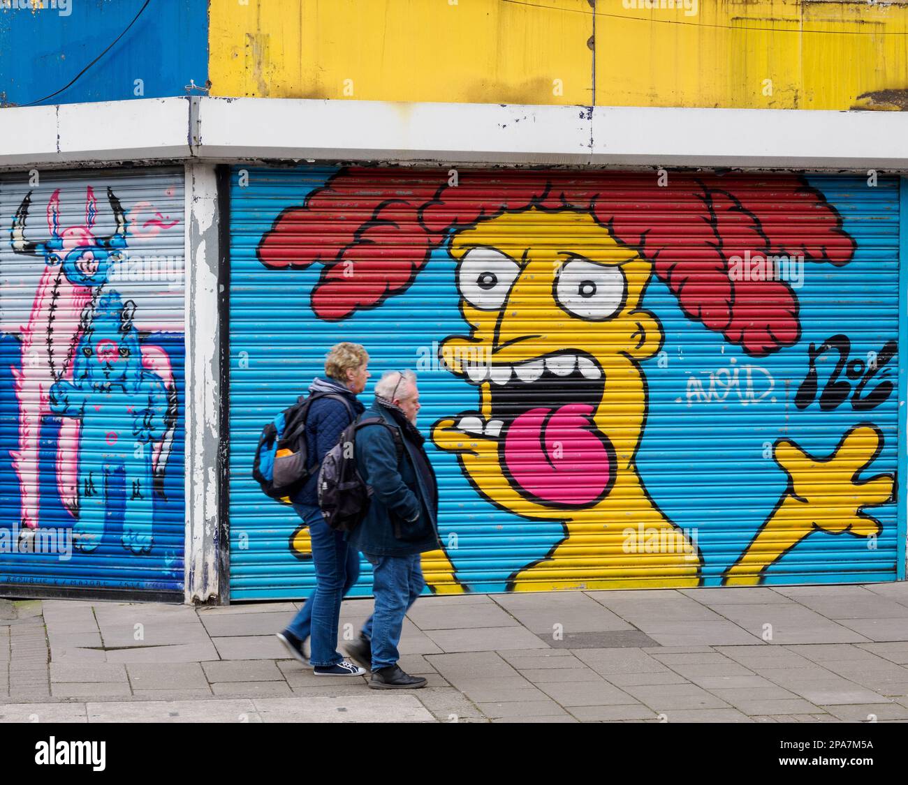 Un uomo e una donna che passano tranquillamente sorprendenti graffiti sulle persiane di un negozio a Bristol UK Foto Stock