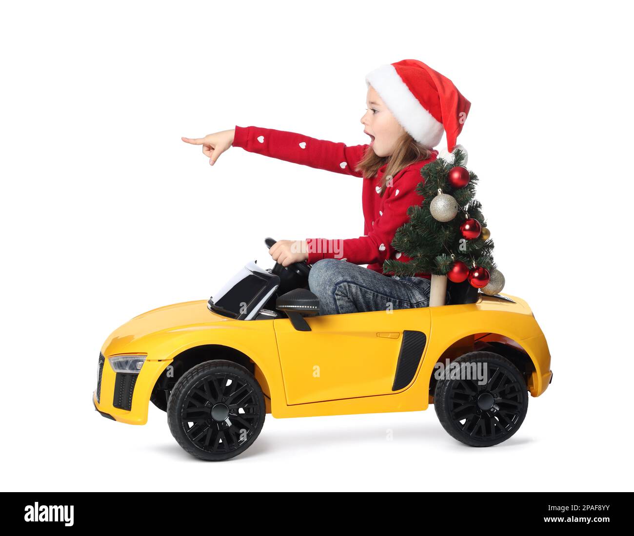 Carina bambina in cappello di Santa con albero di Natale che guida l'auto giocattolo elettrica dei bambini su sfondo bianco Foto Stock