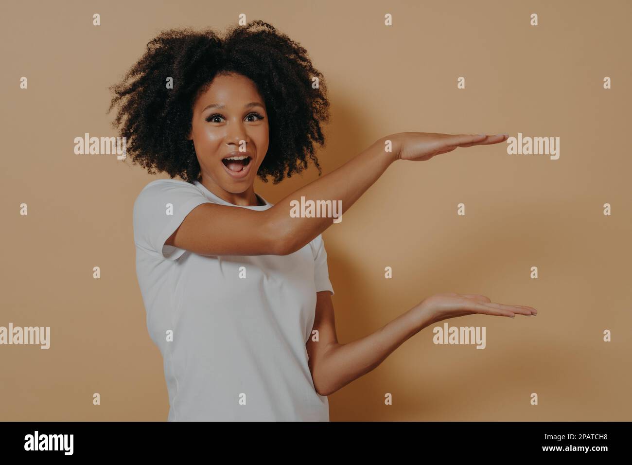 Eccitato Afro americano bella giovane donna con capelli ricci neri che mostra con le mani esempio di formato per la pubblicità, isolato su scuro beige backgro Foto Stock