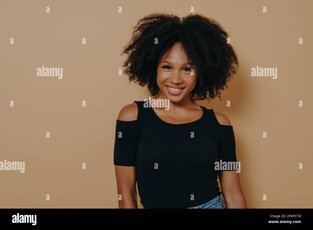 Sorridente afro donna americana con capelli ricci in piedi su sfondo beige, ha felice e positiva espressione del viso, giovane africana femmina in casual Foto Stock