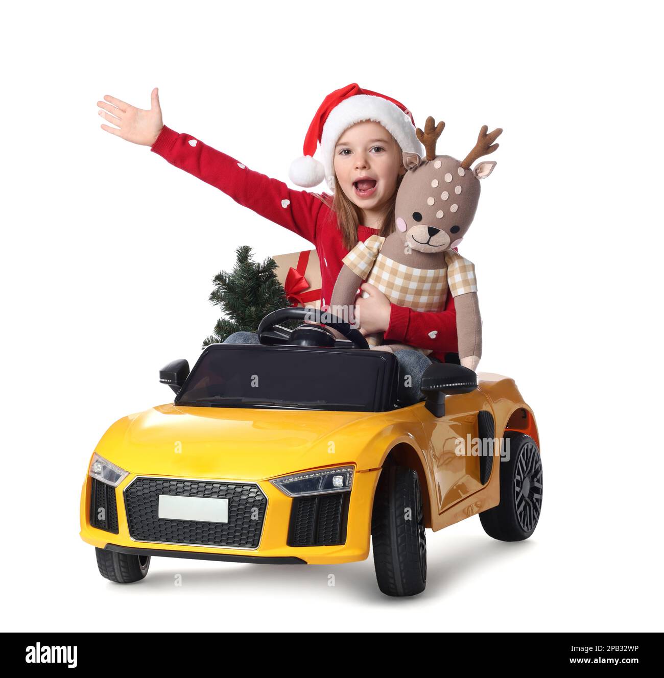 Carina bambina in cappello di Santa con albero di Natale e capriolo giocattolo guida l'auto dei bambini su sfondo bianco Foto Stock
