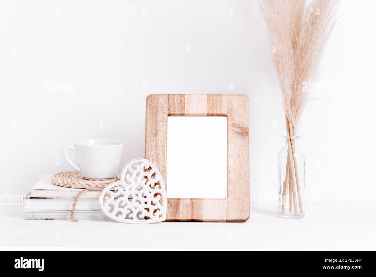 Cornice in legno mockup, libri, caffè, cuore scolpito decor e pampas erba in vaso su tavolo bianco. Spazio di copia. Foto Stock