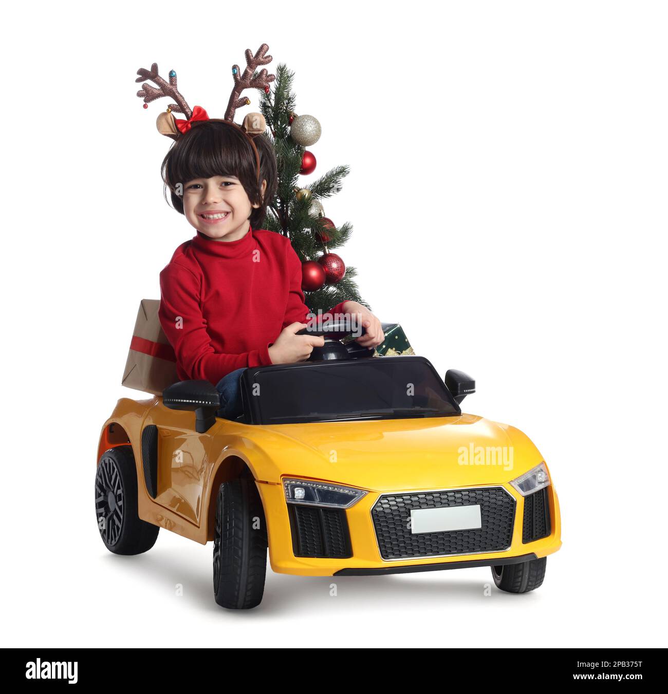 Carino ragazzino con albero di Natale e scatola regalo che guida l'auto giocattolo elettrica dei bambini su sfondo bianco Foto Stock