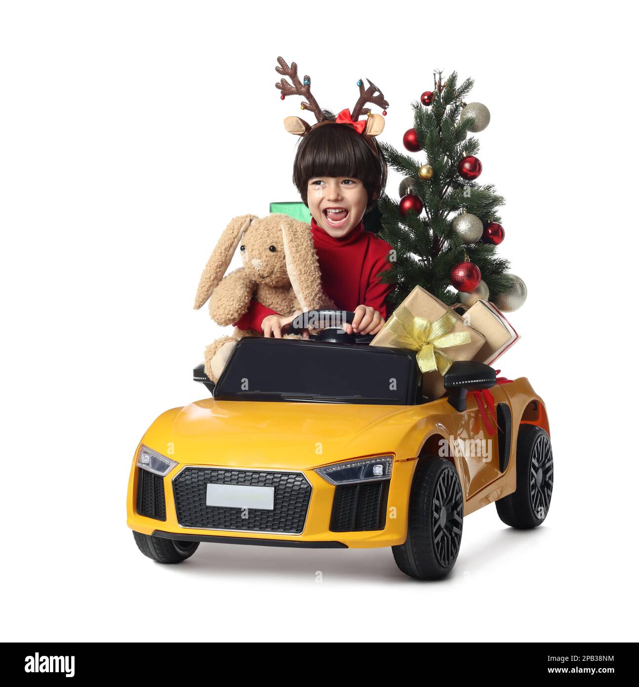 Carino ragazzino con albero di Natale, coniglio giocattolo e scatole regalo che guidano l'auto dei bambini su sfondo bianco Foto Stock