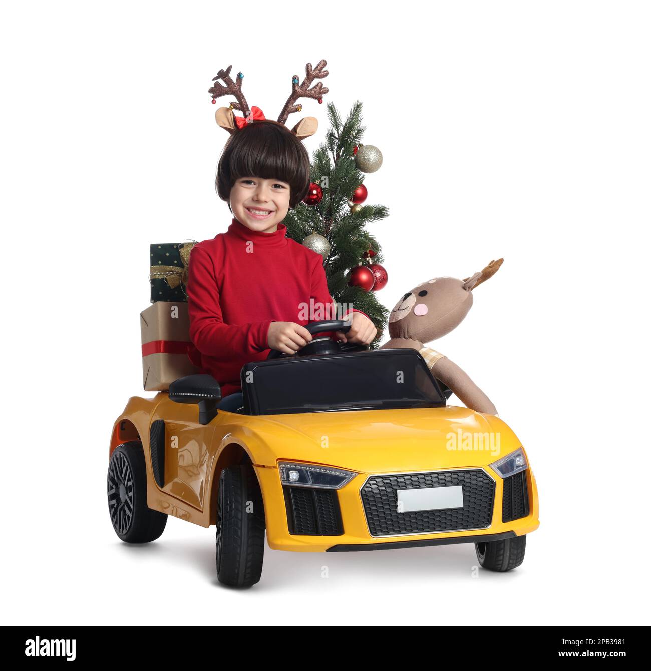 Carino bambino con albero di Natale, giocattolo cervi e scatole regalo che guidano l'auto dei bambini su sfondo bianco Foto Stock