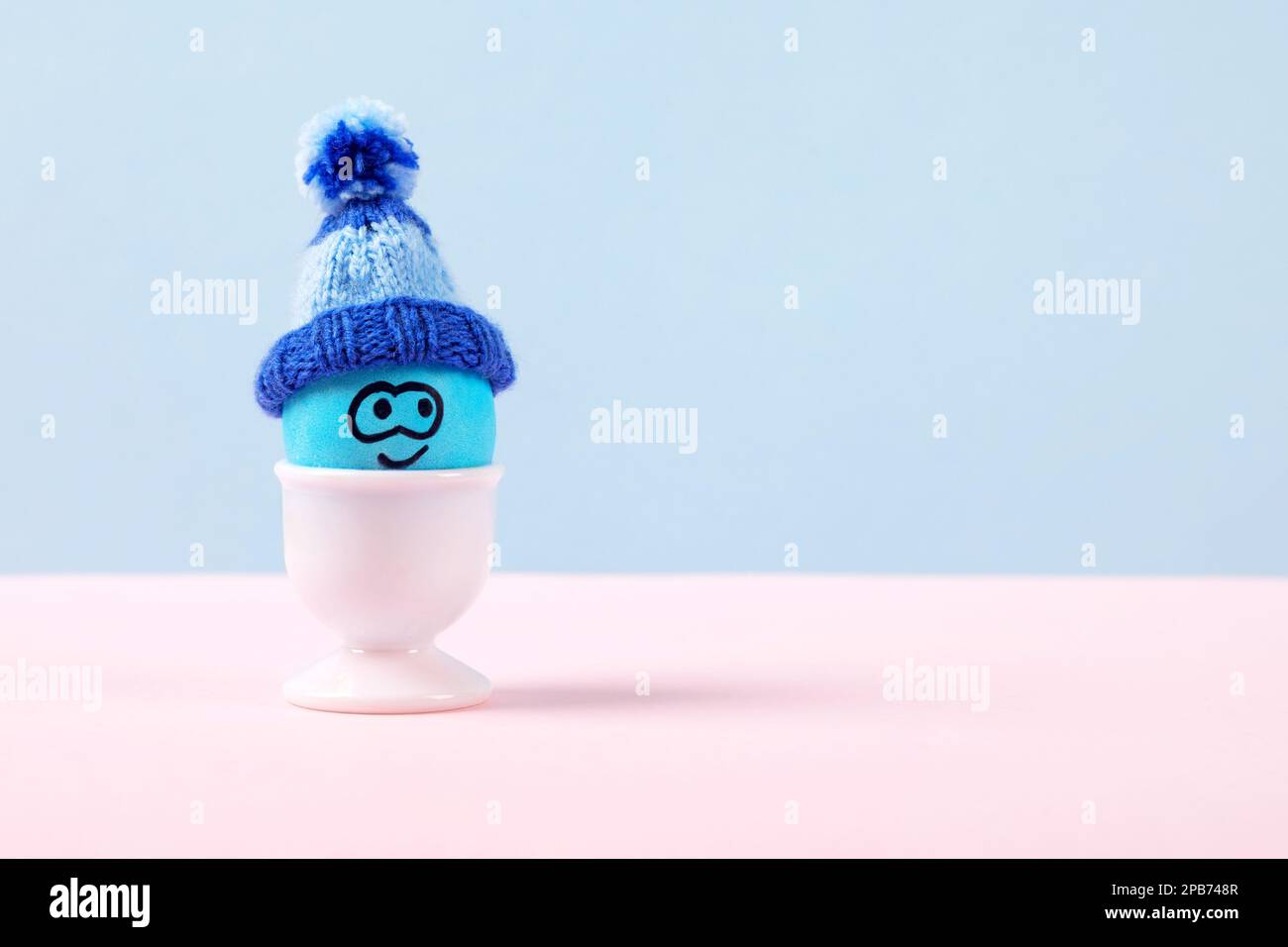 Un uovo di Pasqua blu con volto divertente e cappello a crochet in bianco si trova su un tavolo rosa. Buon concetto di Pasqua. Biglietto d'auguri. Foto Stock