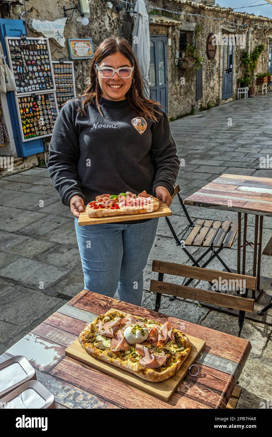 Un ristoratore del piccolo villaggio balneare di Marzamemi porta pizze farcite fatte in casa ai turisti seduti ai tavoli sotto il sole primaverile. Sicilia Foto Stock