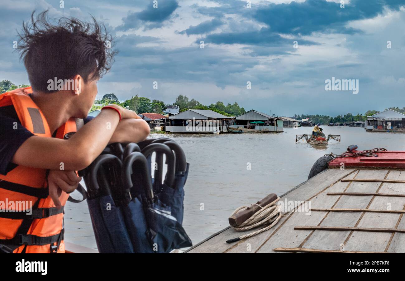 Un giovane su un tender di legno guarda una tradizionale barca da pesca accanto alle fattorie di pesce sul fiume Mekong vicino a Tan Chau in Vietnam. Foto Stock