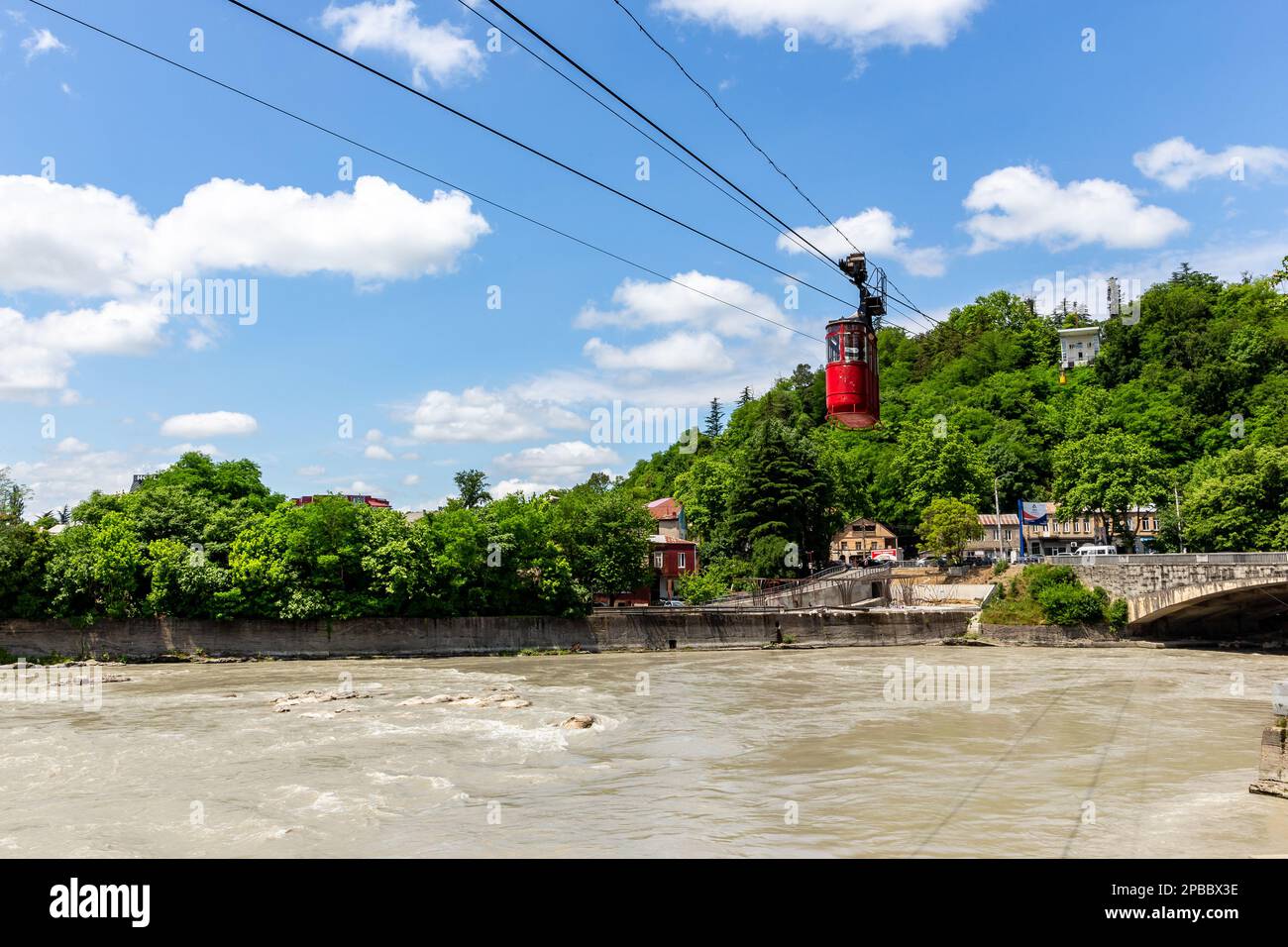 Kutaisi, Georgia, 04.06.21. Funivia rossa Kutaisi sul fiume Rioni, lussureggianti alberi verdi, case residenziali e stazione superiore sullo sfondo. Foto Stock