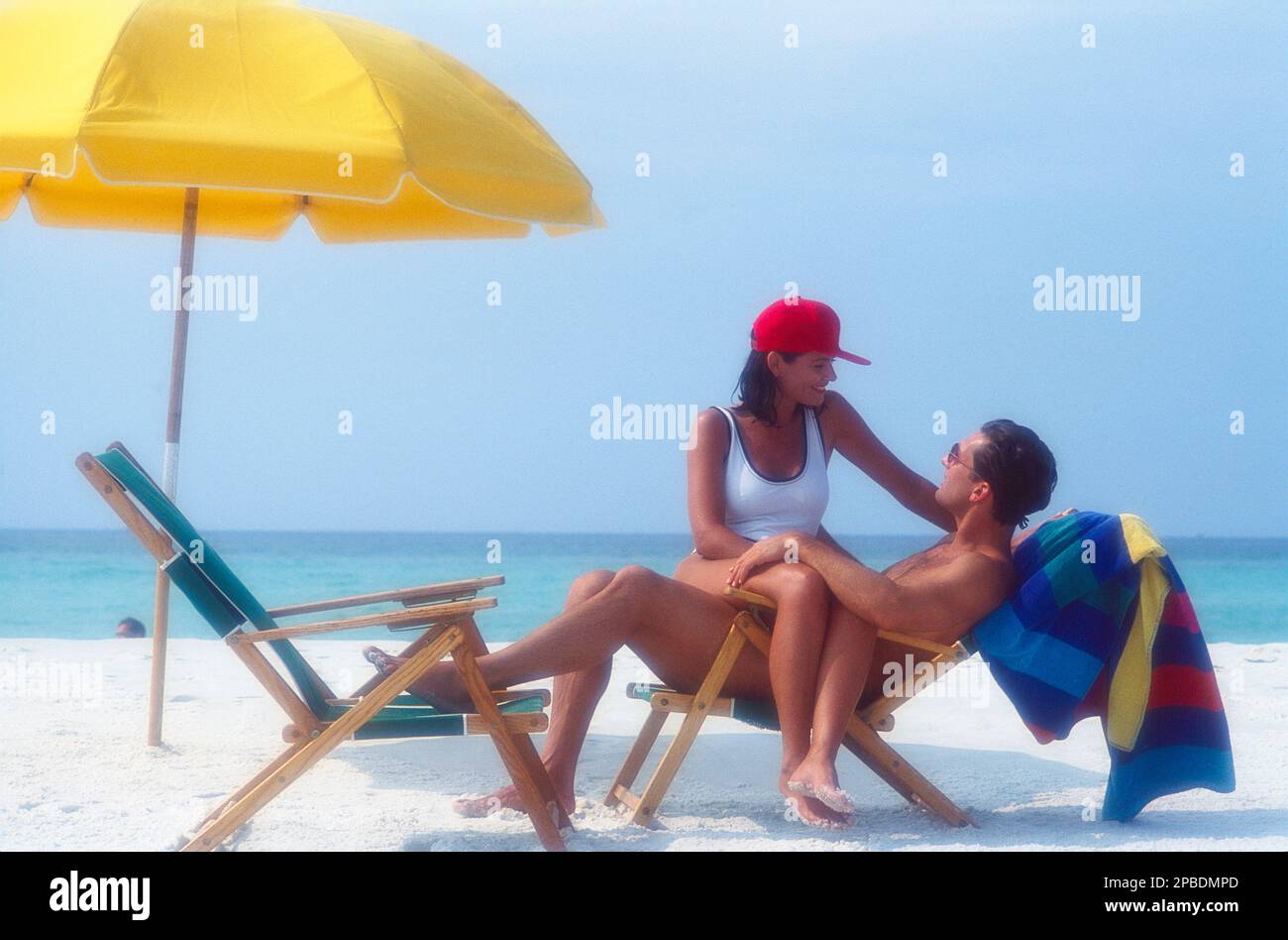 Una giovane coppia caucasica sulla spiaggia dove una donna è seduta in grembo dell'uomo mentre parlano con le sedie a sdraio e l'ombrellone nella sabbia dell'oceano Foto Stock