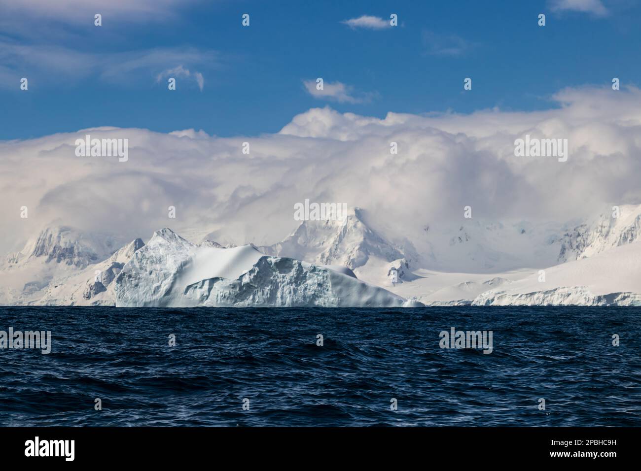Vista sull'oceano della costa della penisola antartica. Montagne con neve e ghiaccio, sotto uno strato spesso di nuvole. Oceano blu scuro in primo piano; cielo blu A. Foto Stock