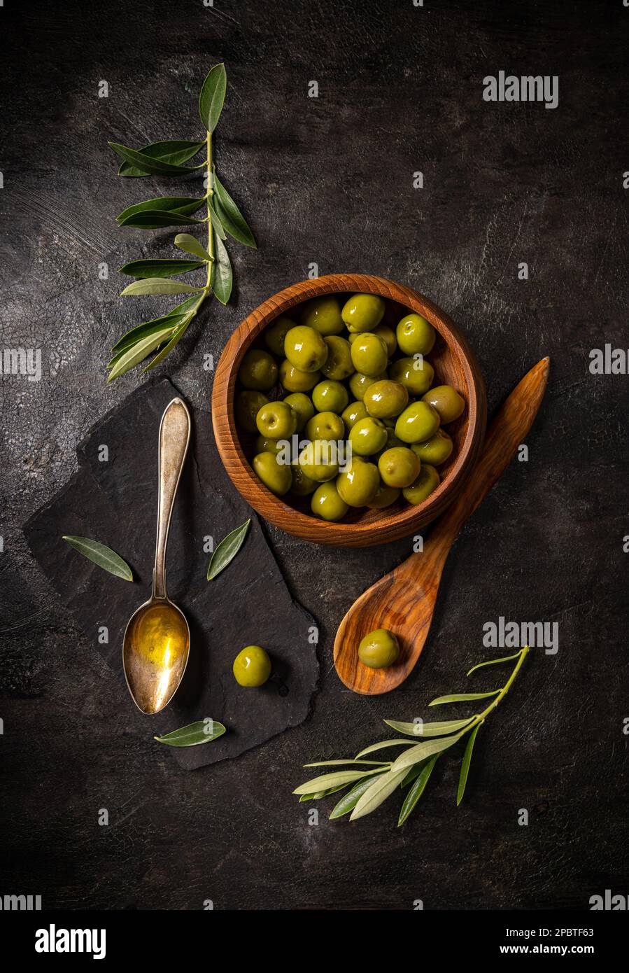 Cucchiaio di olio d'oliva con ramoscello d'oliva fresco e olive su sfondo nero ardesia, vista dall'alto Foto Stock