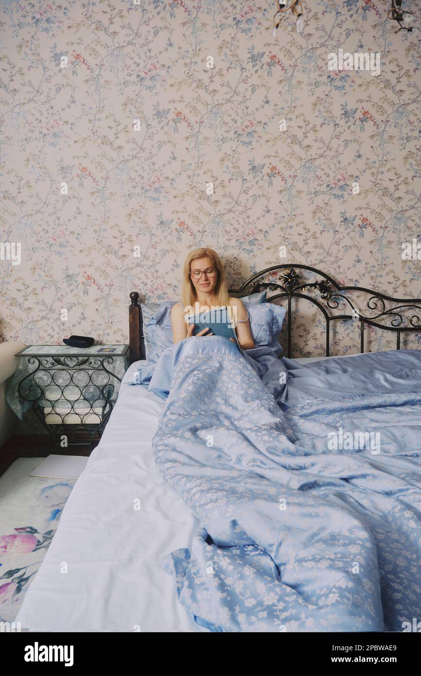 Donna di mezza età che si allunga a letto svegliandosi da sola, tenendo un tablet Foto Stock