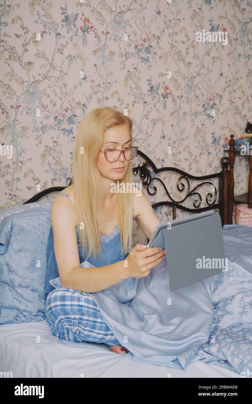 Donna di mezza età che si allunga a letto svegliandosi da sola, tenendo un tablet Foto Stock