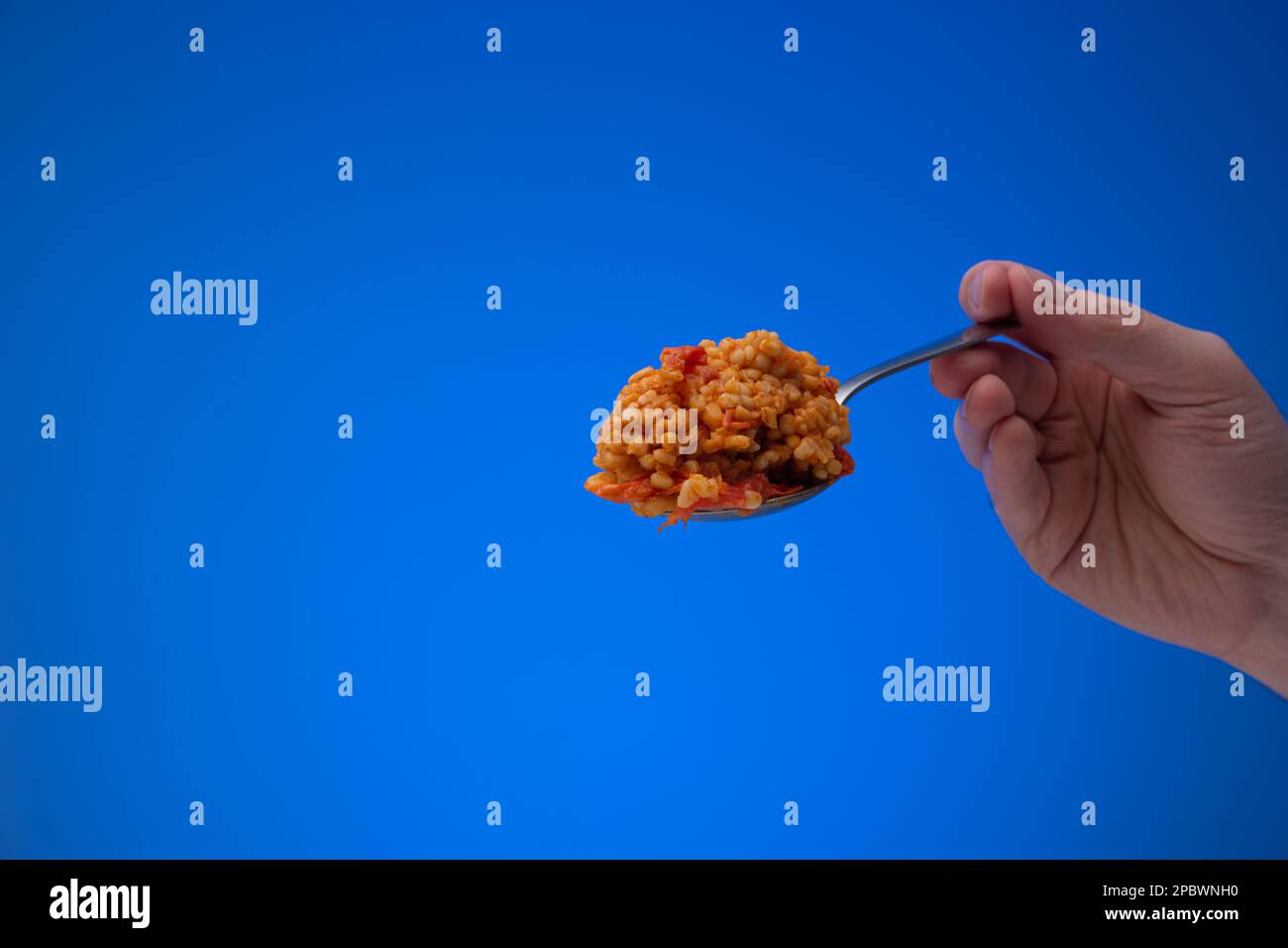 Bulgur cotto con pomodori a dadini su un cucchiaio d'argento tenuto da mano maschio caucasica. Primo piano ripresa in studio, isolata su sfondo blu. Foto Stock