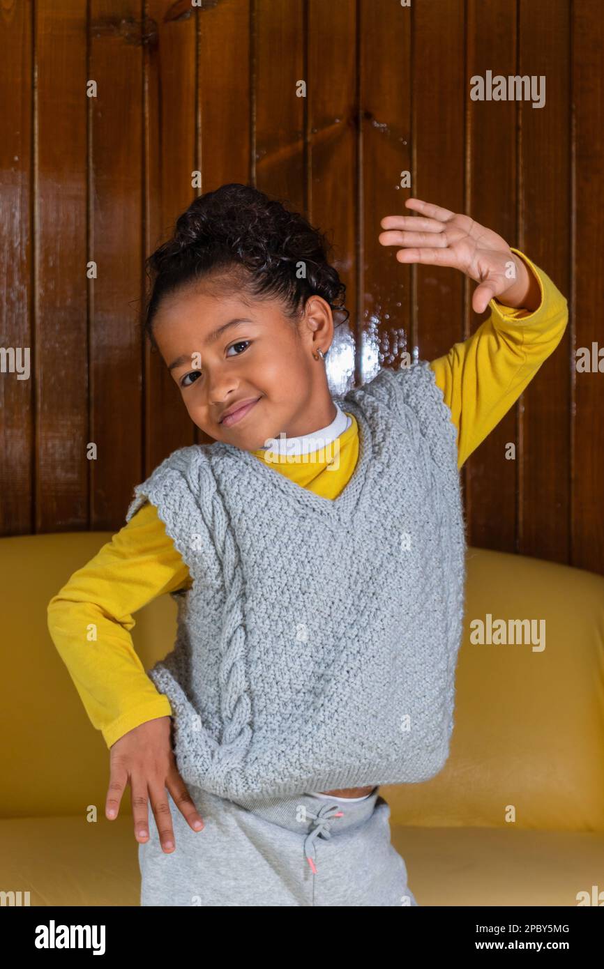 Sorridente ragazza afroamericana prescolare con capelli ricci neri che posano con mano sulla vita vicino al divano giallo e parete di legno mentre guarda la macchina fotografica Foto Stock