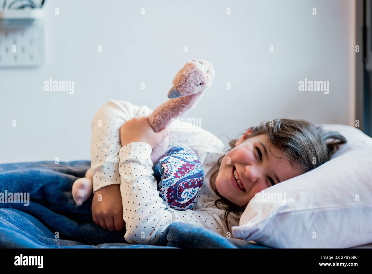 Ragazza allegra in pigiama sdraiata su un morbido cuscino sotto la pedina blu e pigiante uccello giocattolo morbido sguazzare guardando la macchina fotografica prima di andare a dormire Foto Stock