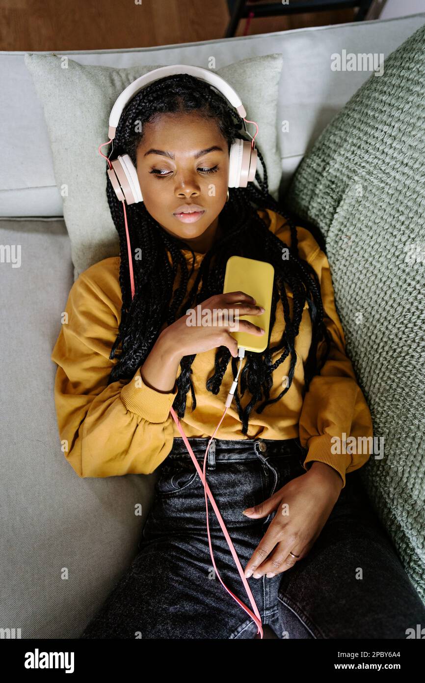 Dall'alto della giovane afro-americana amena non emotiva in abiti casual con trecce sdraiate su un comodo divano e navigare in internet su smartphone whi Foto Stock